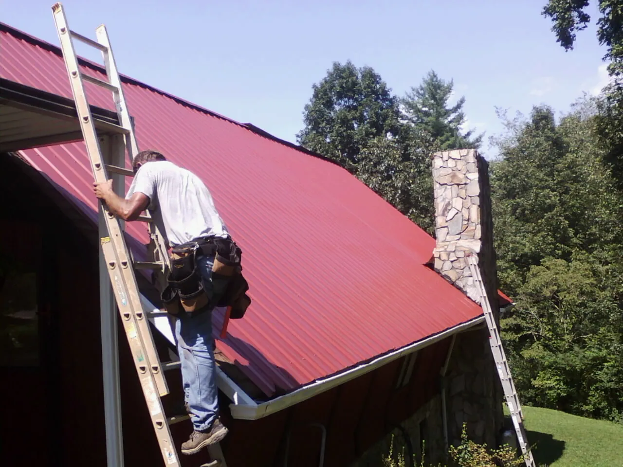 Roofer on a ladder beside a red metal roof and stone chimney — Soffit & Fascia Repair by Roofing Brothers in Waterman